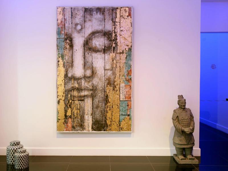 Wandbild mit Gesicht in Erdtönen, daneben Skulptur und schwarz-weiße Vase auf hellem Boden.