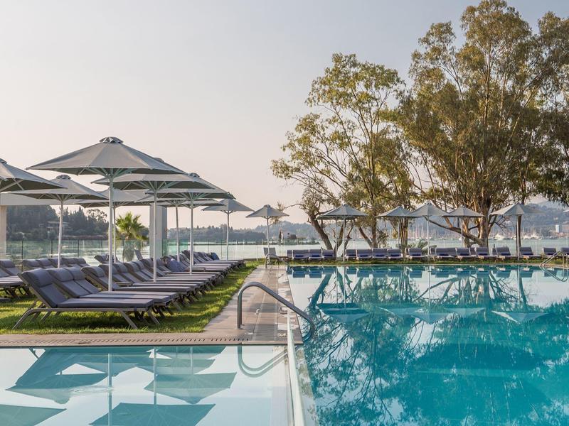 Schwimmbad mit blauer Wasserfläche, Liegestühlen und Sonnenschirmen neben Bäumen am Seeufer.
