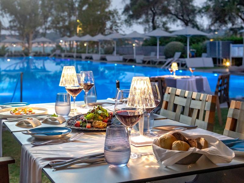 Elegantes Abendessen mit beleuchteten Lampen, Wein und Speisen am Tisch neben blau beleuchtetem Pool.