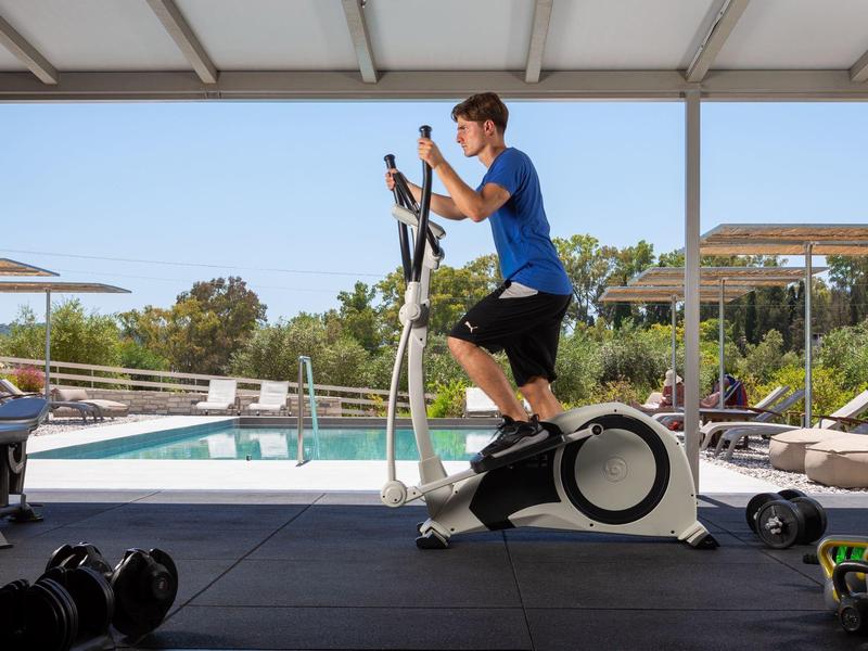 Junge Person trainiert auf Ellipsentrainer neben dem Pool in sonnigem Outdoor-Fitnessbereich.