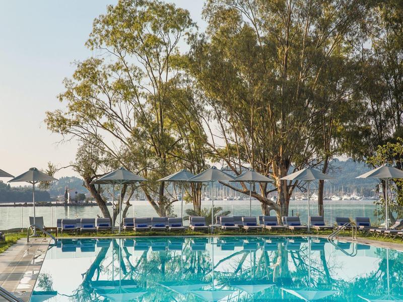 Ein klarer blauer Pool mit Liegestühlen und Sonnenschirmen, umgeben von Bäumen am Wasser.