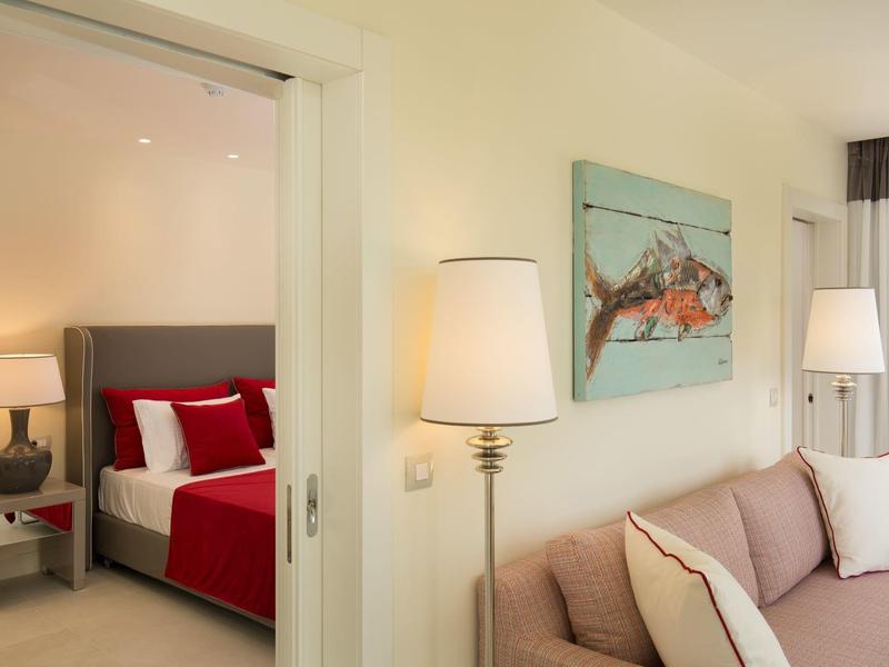 Helles Wohnzimmer mit beigem Sofa, Stehlampe und Schlafzimmer mit rotem Bett im Hintergrund.