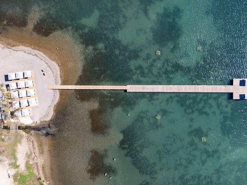 Langer Steg führt von Strand mit Liegen ins klares, türkis-blaues Meer. Sand und Felsen am Ufer.