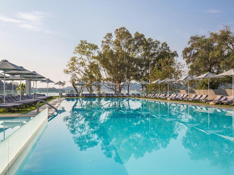 Großer, ruhiger Pool mit blauer Wasserfläche, umgeben von Liegestühlen und Bäumen im Hintergrund.