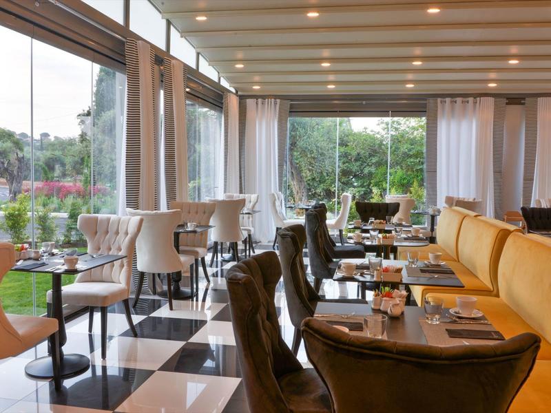 Innenraum eines Restaurants mit großen Fenstern, schwarzen Stühlen, gelben Sofas und schwarz-weißem Boden.