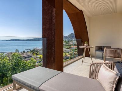 Balkon mit Lounge-Möbeln, Holzsäulen und Blick auf das Meer und grüne Hügel im Hintergrund.
