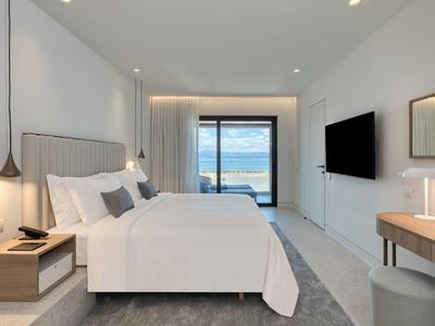 Modernes Schlafzimmer mit großem weißen Bett, Nachttisch, Fernseher und Meerblick durch Fenstertür.
