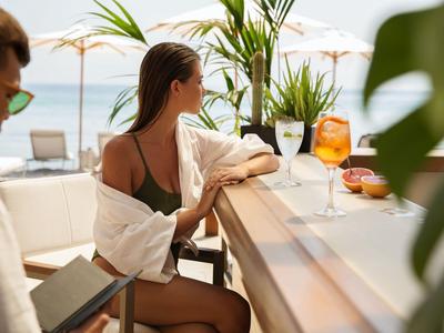 Frau in weißem Hemd sitzt am Bar-Tresen mit Blick auf den Pool und das Meer.