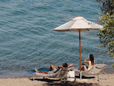 Zwei Personen entspannen auf Liegestühlen unter einem Sonnenschirm am Strand neben blauem Wasser.