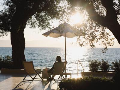 Person entspannt sich auf Liegestuhl am Meer mit Sonnenschirm im Schatten großer Bäume.
