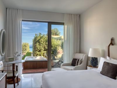 Helles Hotelzimmer mit großem Bett, Sitzbereich und Blick auf einen Garten und das Meer.