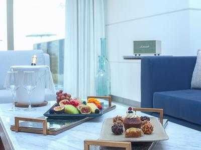 Moderner Hotelraum mit blauer Couch, Obstteller und Dessert auf Tischen vor großen Fenstern.