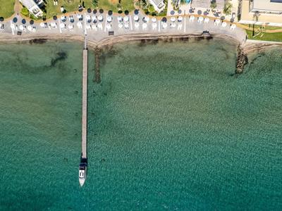 Luftaufnahme eines langen Stegs mit einem Boot an klarem, türkisem Wasser nahe einer belebten Strandpromenade.