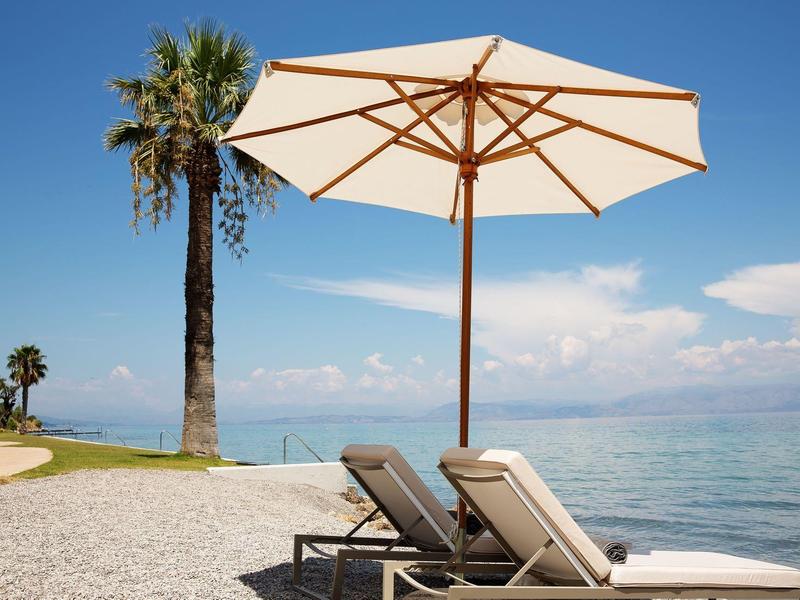 Zwei Liegestühle mit weißem Sonnenschirm am Kiesstrand, Palmen und ruhiges Meer im Hintergrund.