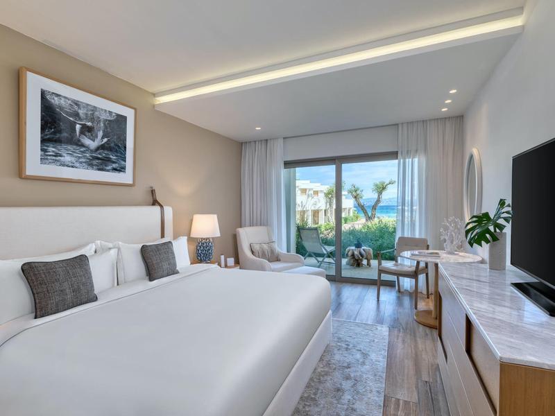 Modernes Hotelzimmer mit großem Bett, TV, Sessel und Blick auf Garten und Meer durch große Fenster.