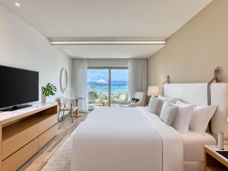 Helles Schlafzimmer mit großem Bett, Fernseher auf Kommode und Meerblick durch Fenster.