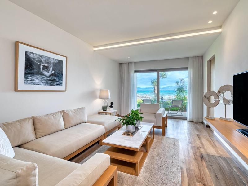 Helles Wohnzimmer mit beigem Sofa, Holzmöbeln, Teppich und großem Fenster mit Blick nach draußen.