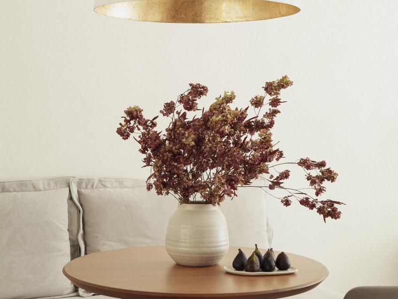 Moderner Wohnbereich mit rundem Holztisch, Vase mit getrockneten Zweigen und einer weißen Hängeleuchte.