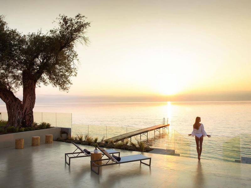 Frau steht auf Terrasse mit Liegestuhl, Baum links, Blick auf Sonnenuntergang über ruhigem Meer.