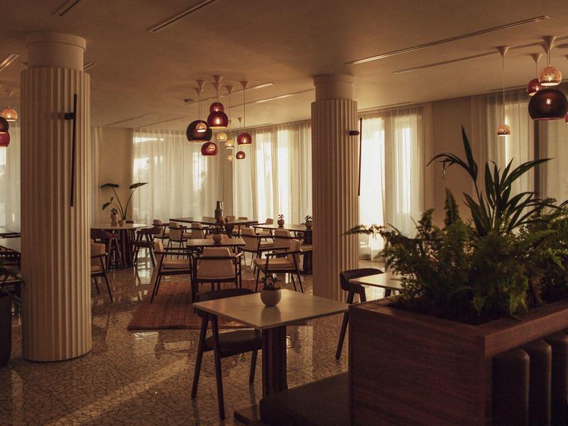 Helles Restaurant mit weißen Säulen, Holztischen, Stühlen und großen Pflanzenarrangements.