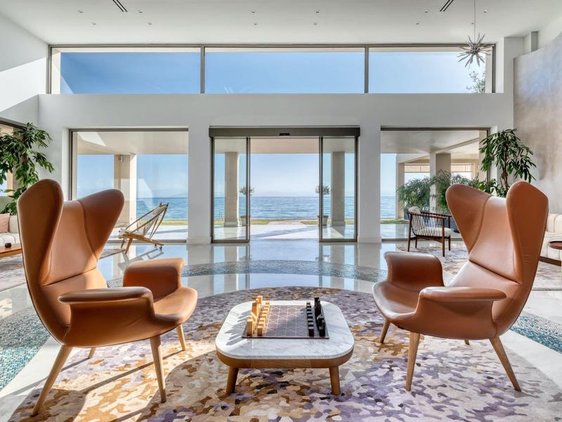 Helles Wohnzimmer mit bodentiefen Fenstern, beigen Ledersesseln und Meerblick im Hintergrund.