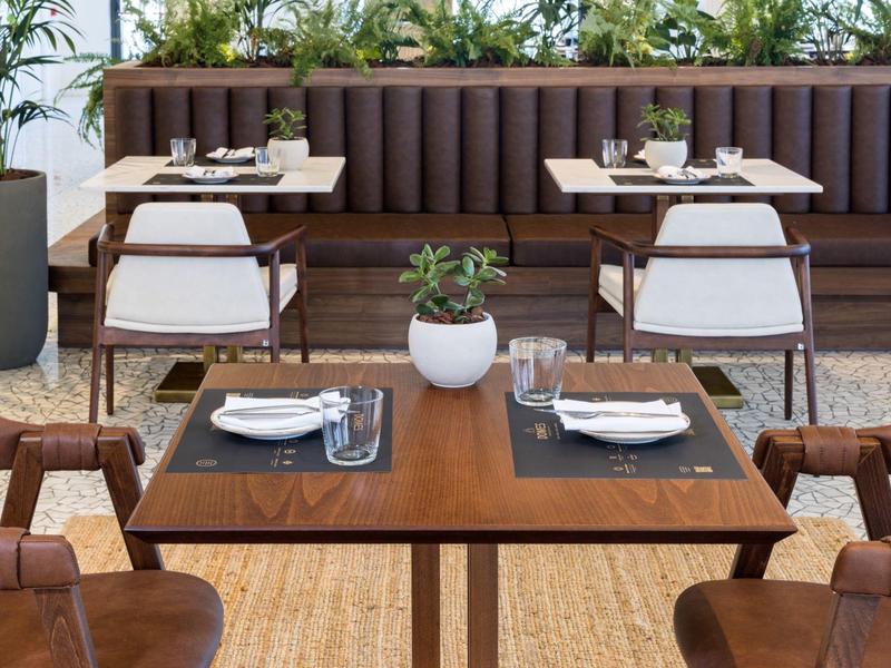 Moderner Restaurantbereich mit braunen Holztischen, gepolsterten Stühlen und grünen Pflanzen.