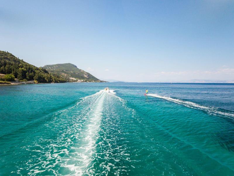Klares türkisfarbenes Meer mit Bootsspur und bewaldeten Hügeln im Hintergrund unter blauem Himmel.