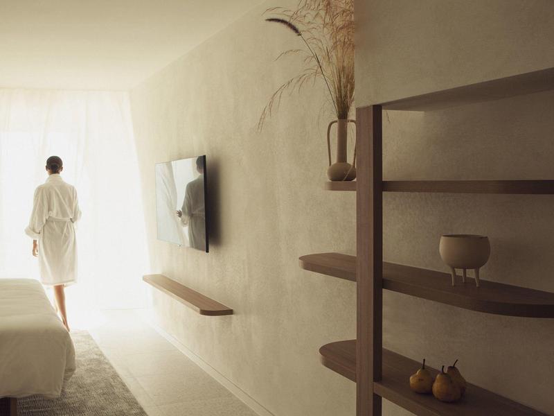 Helles Schlafzimmer mit weißem Bett, Wandregalen, Fernseher und Person im Bademantel am Fenster.