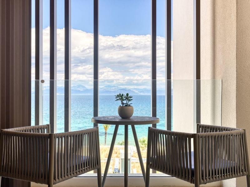Zwei graue Stühle und ein Tisch mit Pflanze vor einem großen Fenster mit Blick auf Meer und Himmel.