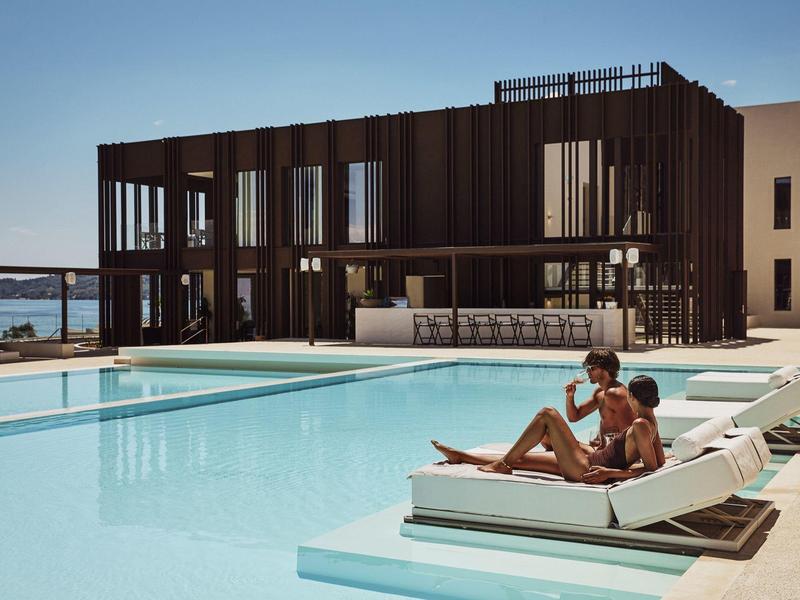 Moderner Poolbereich mit Liegestühlen, zwei entspannten Personen und einem dunklen Gebäude im Hintergrund.