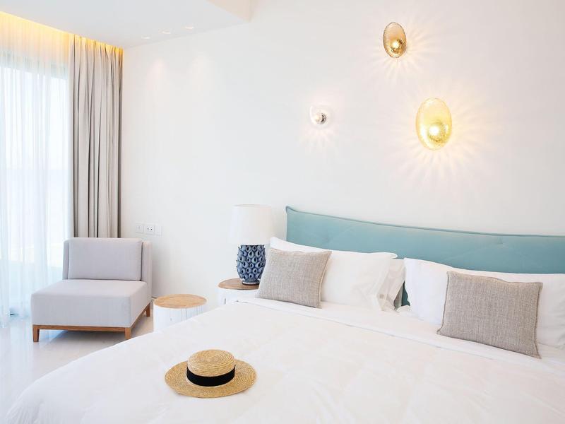 Helles Schlafzimmer mit großem Bett, blauer Rückenlehne, grauen Kissen, Sessel und Wandlampen.