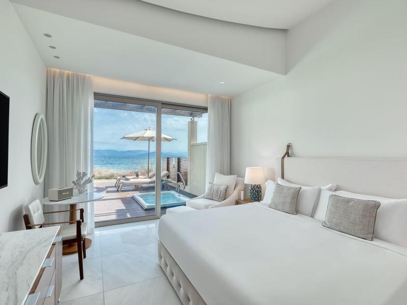 Hell eingerichtetes Schlafzimmer mit großem Bett, Fernseher, Schreibtisch und Meerblick durch Glasschiebetür.