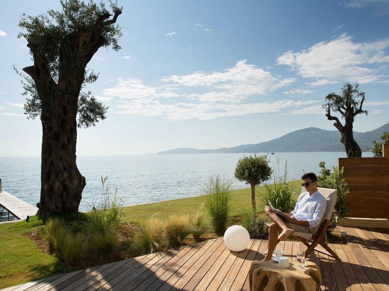 Mann sitzt auf Holzterrasse am See, umgeben von Bäumen und Bergen im Hintergrund.