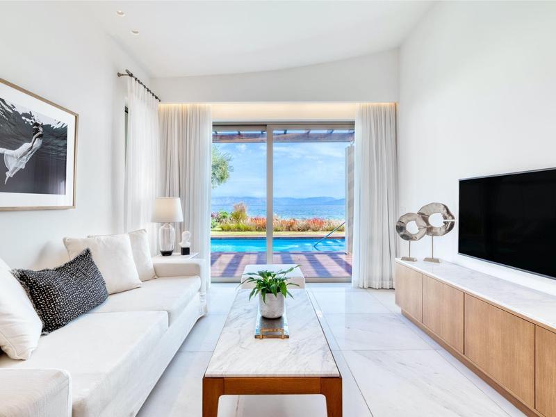 Helles Wohnzimmer mit weißem Sofa, Holztisch, TV-Möbel und Blick auf Pool und Natur draußen.