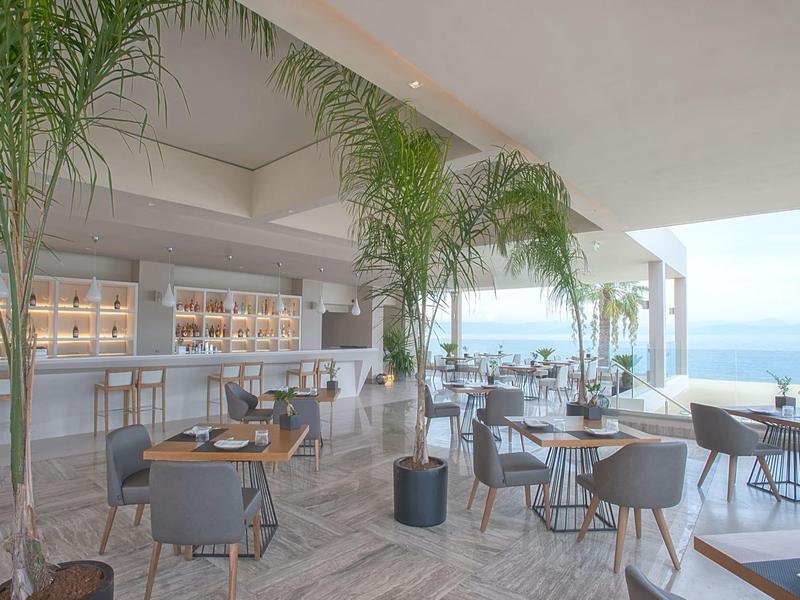 Helle, moderne Strandbar mit Palmen, runden Tischen und Blick auf das Meer.