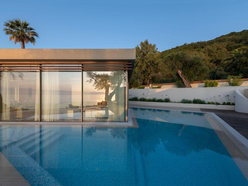 Moderner Pool mit klarem Wasser, Glasfassade und grünen Hügeln im Hintergrund bei blauem Himmel.