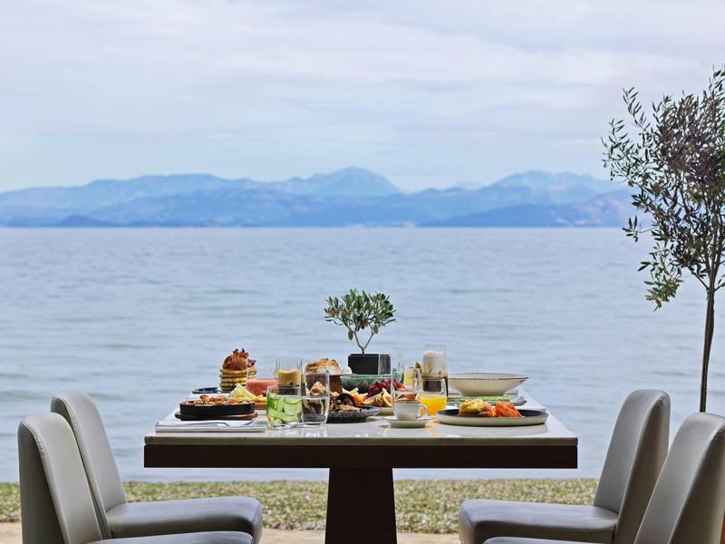 Gedeckter Tisch mit Stühlen am Wasser, umgeben von kleinen Bäumen und Bergen im Hintergrund.