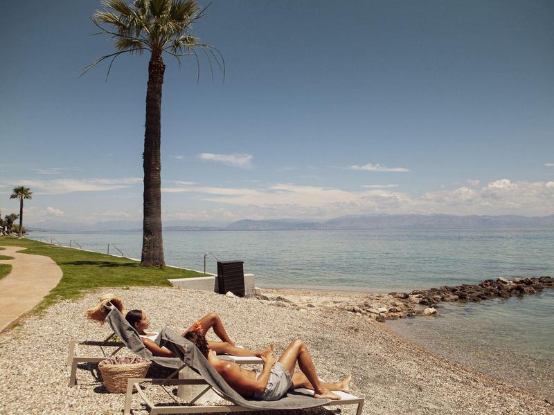 Zwei Personen liegen auf Liegestühlen am Kiesstrand mit Palmen und Blick aufs Meer bei blauem Himmel.