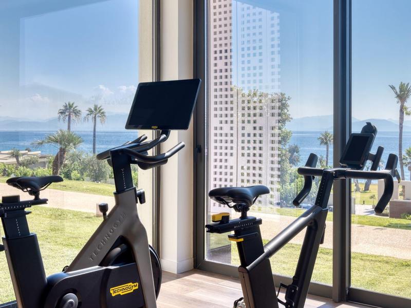 Hell eingerichteter Fitnessraum mit schwarzen Indoor-Bikes vor großen Fenstern und Meerblick.