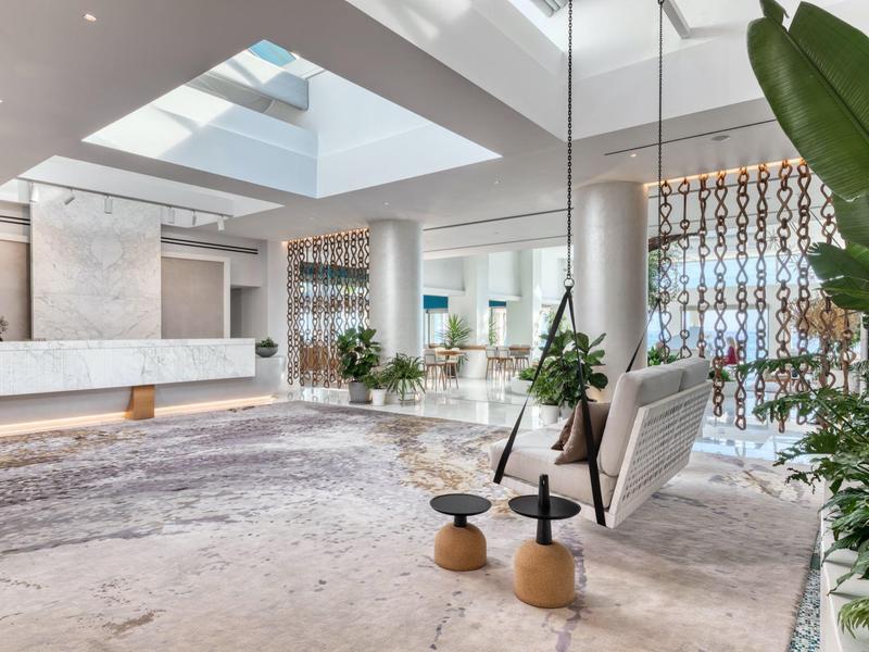 Moderner, heller Innenraum mit großen Fenstern, Pflanzen, Sitzmöbeln und strukturiertem Teppichboden.