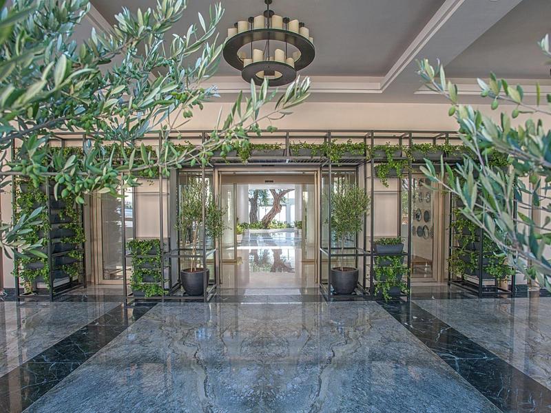 Modernes Hotelfoyer mit Glas-Eingangstüren, glänzendem Marmorfußboden und grünen Pflanzen.