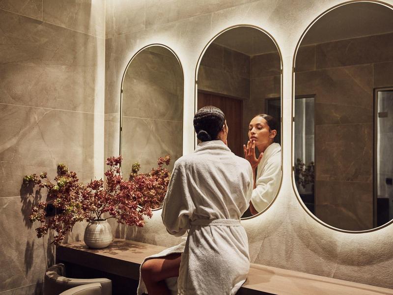 Frau im weißen Bademantel schaut in einen ovalen Spiegel in einem hell beleuchteten Badezimmer.