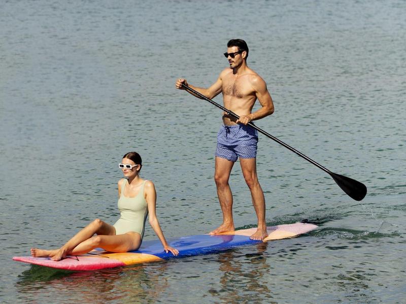Mann steht mit Paddel, Frau sitzt auf buntem Paddleboard auf ruhigem Meer in klarer Sicht.