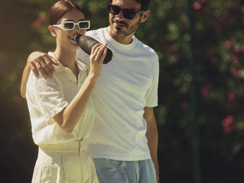 Ein Paar in Freizeitkleidung umarmt sich lachend auf einem Tennisplatz bei Sonnenlicht.