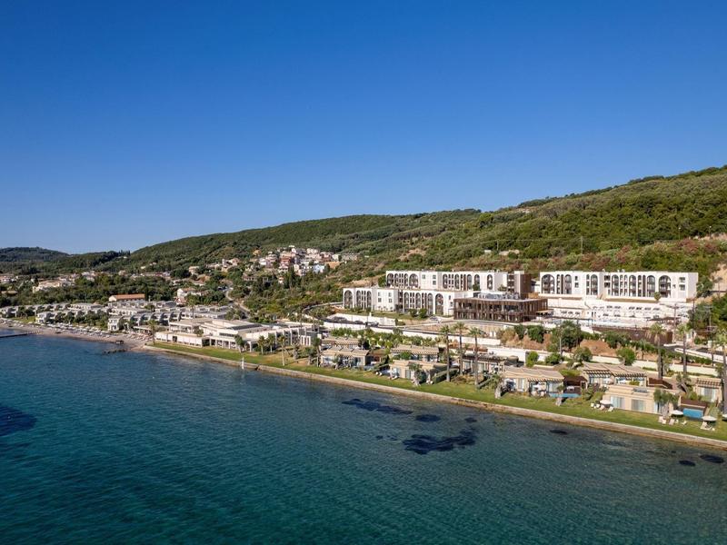 Küste mit klarem blauen Wasser, Gebäuden am Strand und grünen Hügeln im Hintergrund.
