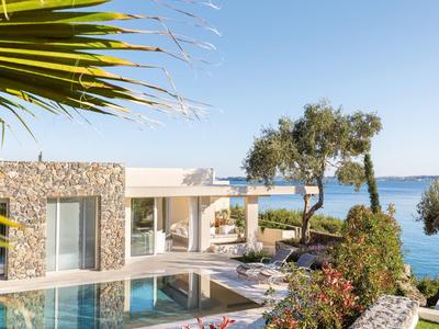 Modernes Haus mit Pool, Steinfassade, Palmen und Blick auf das blaue Meer bei klarem Himmel.