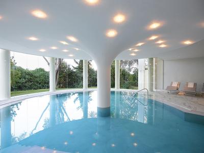 Innenraum mit rundem Pool, weißen Wänden, Säule in der Mitte und großen Fenstern zum Garten.