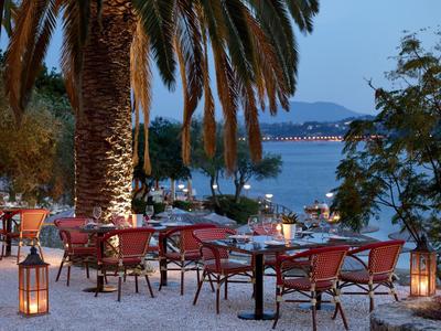 Terrazza serale con tavoli e sedie sotto palme, vista su una baia illuminata.