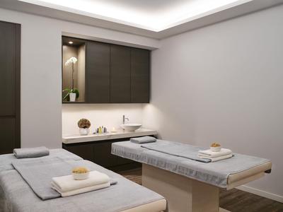 Behandlungsraum mit zwei Liegen, grauen Bezügen, Handtüchern und gedämpftem Deckenlicht.