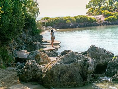 Persona in piedi sulle rocce vicino alla riva di acqua calma con vegetazione verde sullo sfondo.